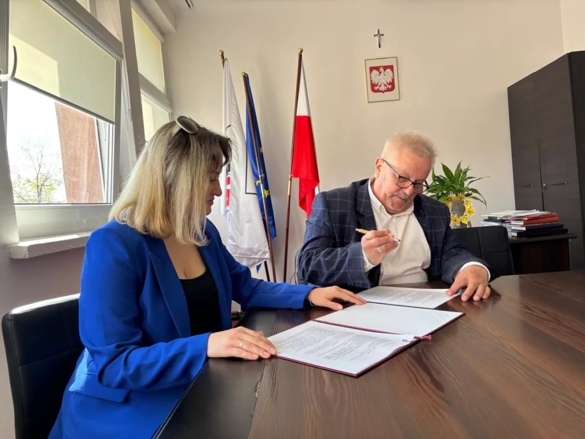 Mężczyzna w okularach, białej koszuli i garniturze, podpisuje przy stole w biurze dokument. Obok siedzi kobieta z jasnymi włosami w niebieskiej marynarce