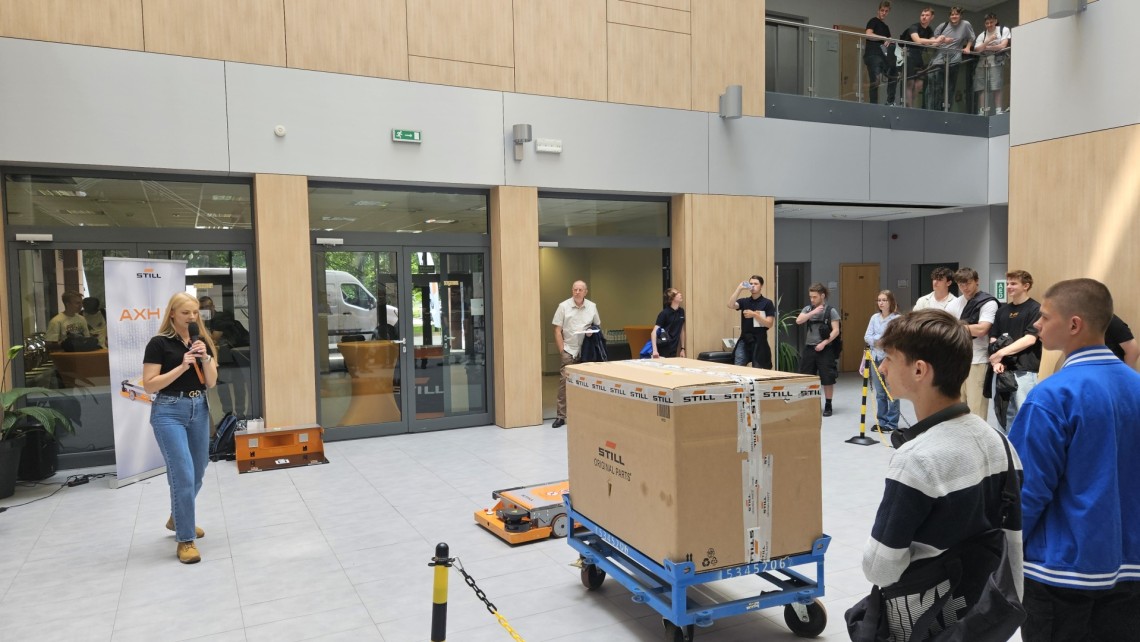 Kobieta z mikrofonem prezentuje autonomicznego robota ciagnącego dużą paczkę, co obserwują młodzi ludzie