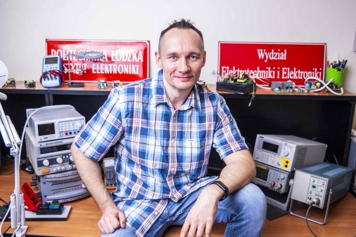 Osobę siedzącą przed biurkiem z różnym sprzętem elektronicznym i urządzeniami. Za osobą znajdują się czerwone tablice, po lewej stronie z napisem POLITECHNIKA ŁÓDZKA WYDZIAŁ ELEKTRONIKI, a znak po prawej  z napisem "Wydział Elektrotechniki i Elektroniki."