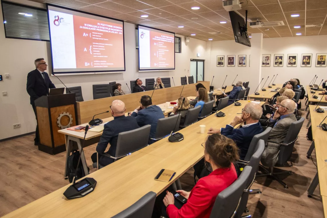 Osoby w sali seminaryjnej słuchają przemawaijącego mężczyzny.