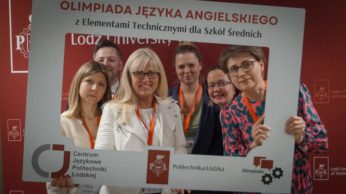Kilka osób ustawiło się w dużej ramce do zdjęcia, na której jest napis Olimpiada Języka Angielskiego