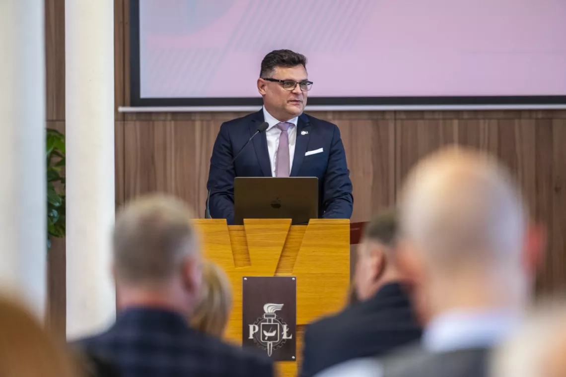 Mężczyzna w garniturze stoi za mównicą i przemawia. Na mównicy leży laptop. Z tyłu widoczny fragment białego ekranu.