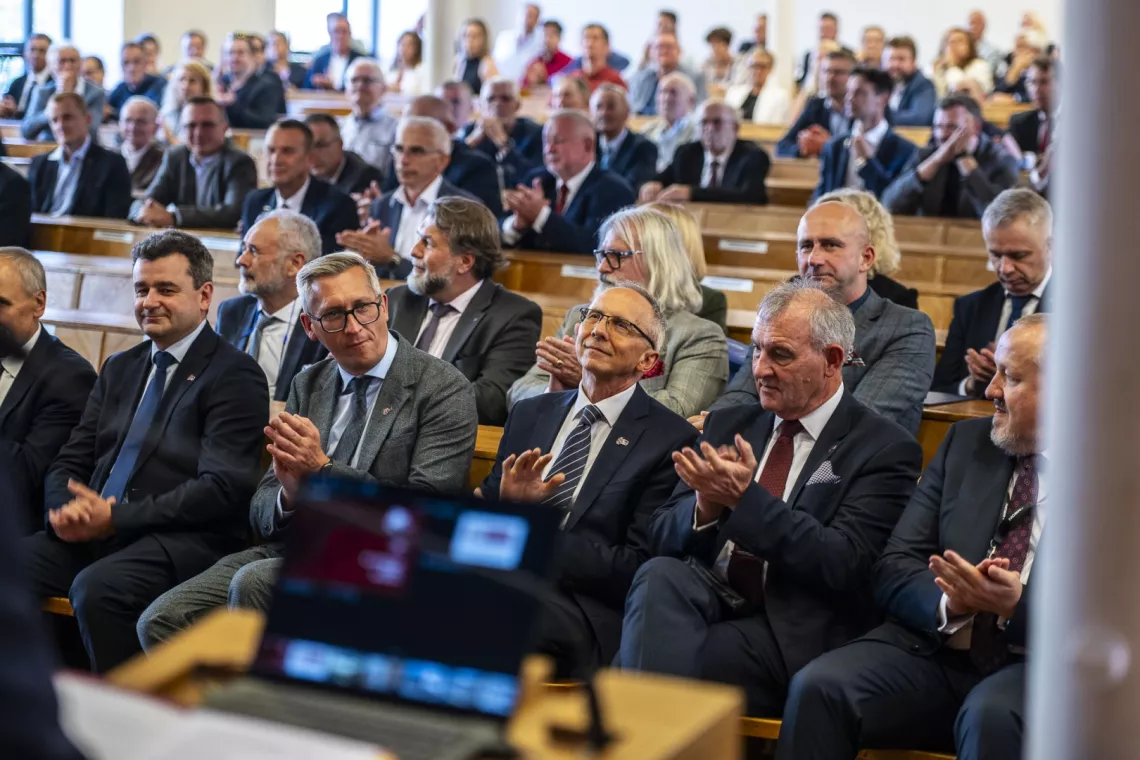 Goście uroczystości biją brawo. Siedzą w ławach w audytorium.