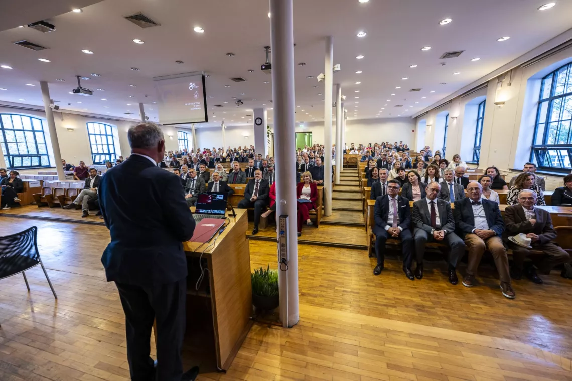 Duże audytorium pełne gości biorących udział w uroczystym spotkaniu. Na pierwszym planie osoba przemawiająca przy mównicy, widoczna od strony pleców
