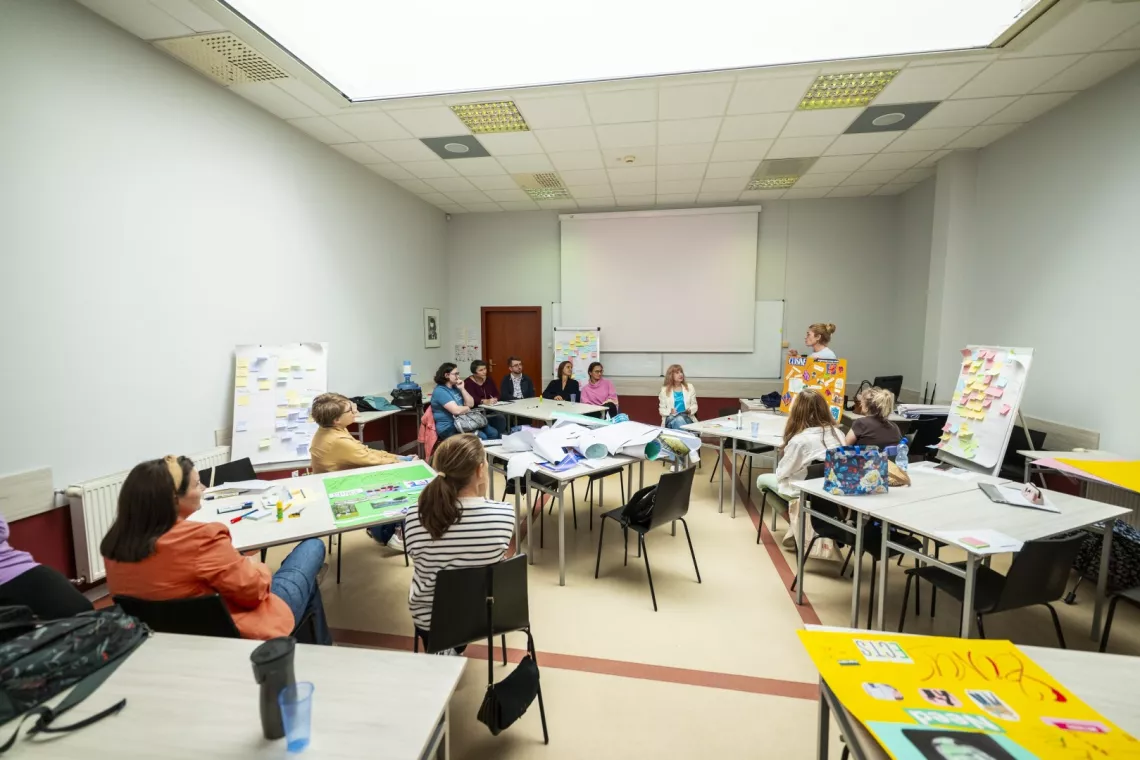 Grupa osób siedzi w sali szkoleniowej, słucha prezentacji prowadzonej przez stojącą przy stole, która trzyma kolorowy plakat