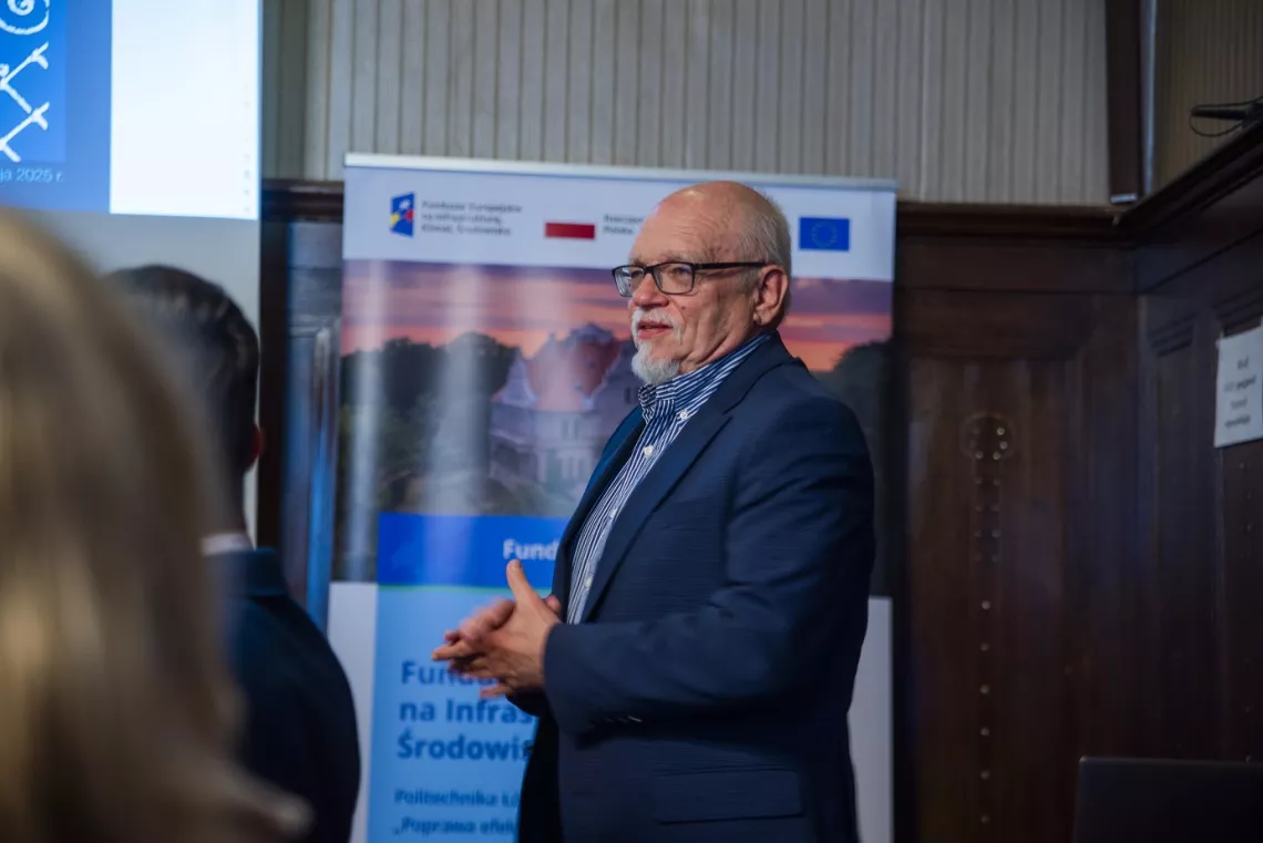 Mężczyzna w okularach i garniturze wygłasza prelekcję, z tyłu widoczny jest baner