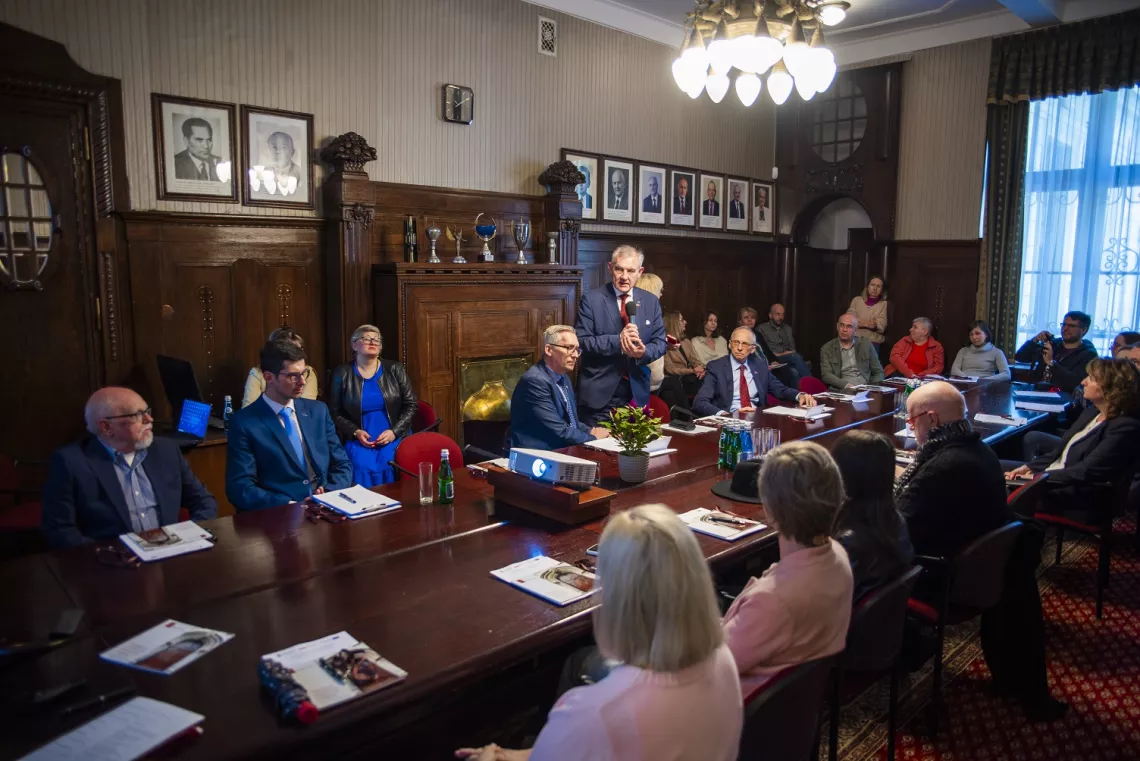 Uczestnicy spotkania siedzą w zabytkowym wnętrzu przy stole, na którym leżą papiery i małe albumy