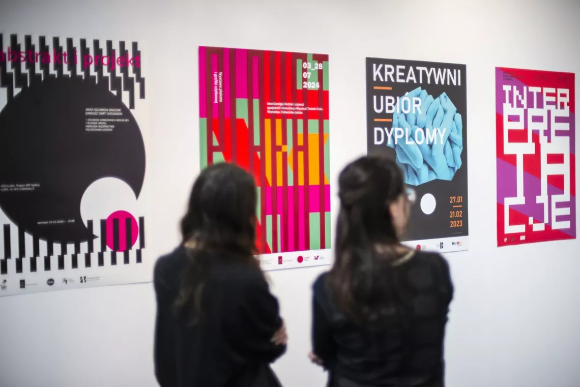 Prace graficzne umieszczone na ścianie oglądają dwie osoby