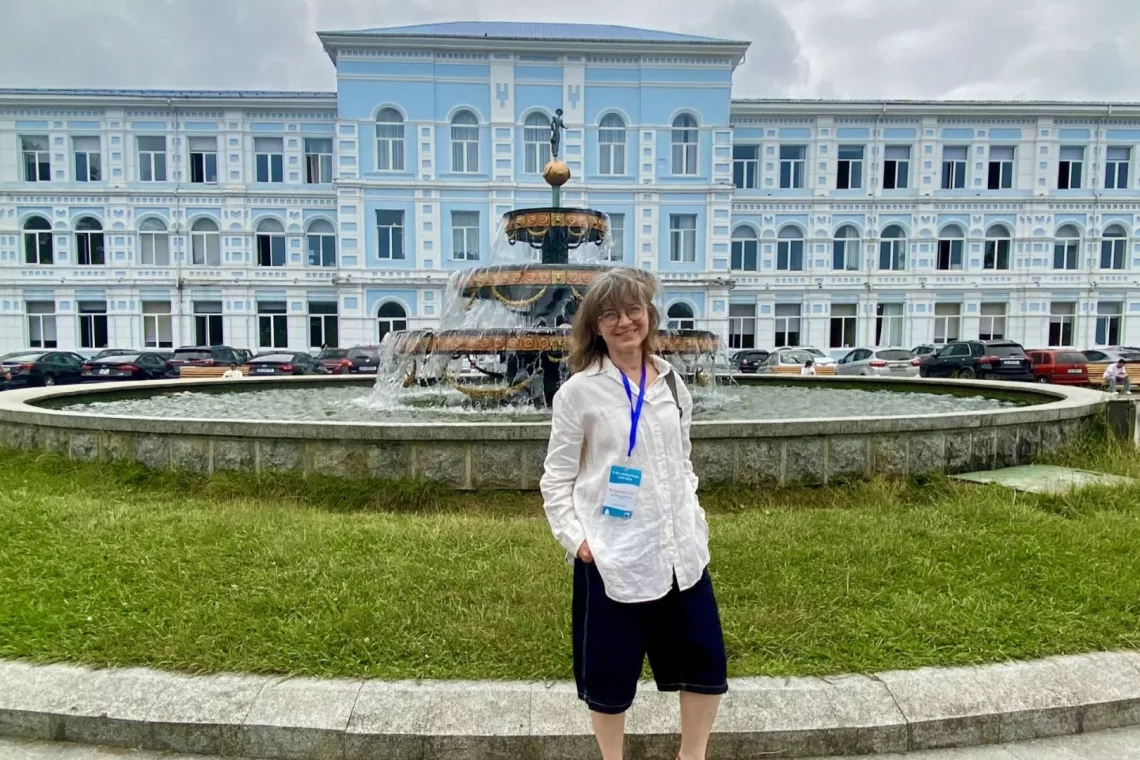 Kobieta w białej koszuli i spodniach stoi przed fontanną, na tle niebieskiego trzypiętrowego budynku 