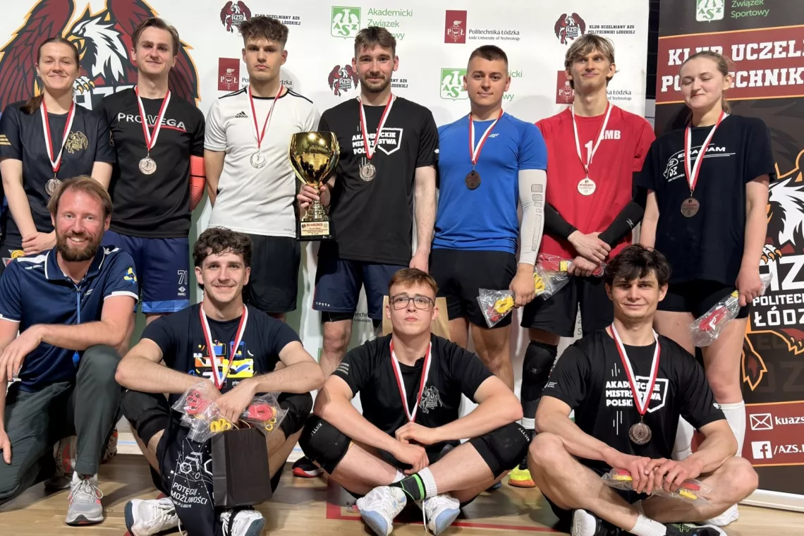 Kilkanaście osób w sportowych strojach z medalami oraz pucharem stoi na tle rollaupu z napisem AZS PŁ 