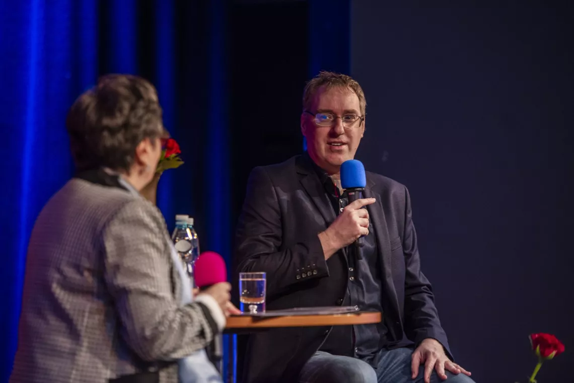 Mężczyzna z mikrofonem udziela wywiadu kobiecie, z którą siedzi przy okrągłym stoliku.