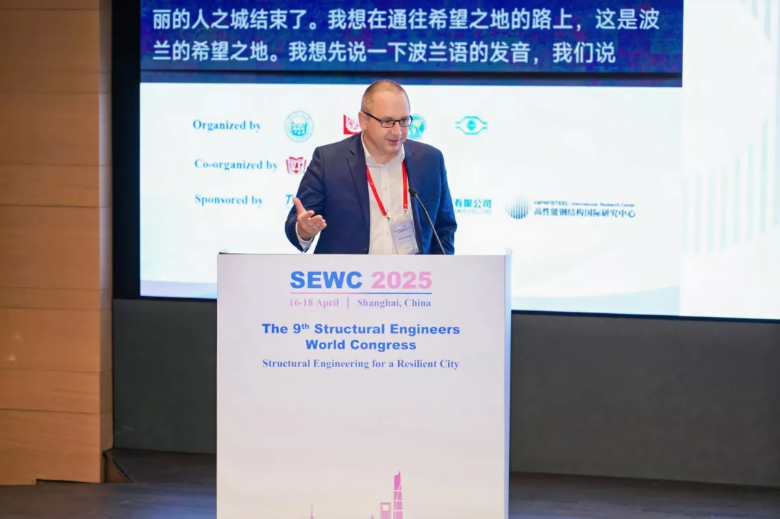 Mężczyzna w garniturze stoi za mównicą i prowadzi wykład. W tle ekran z napisami w języku chińskim.