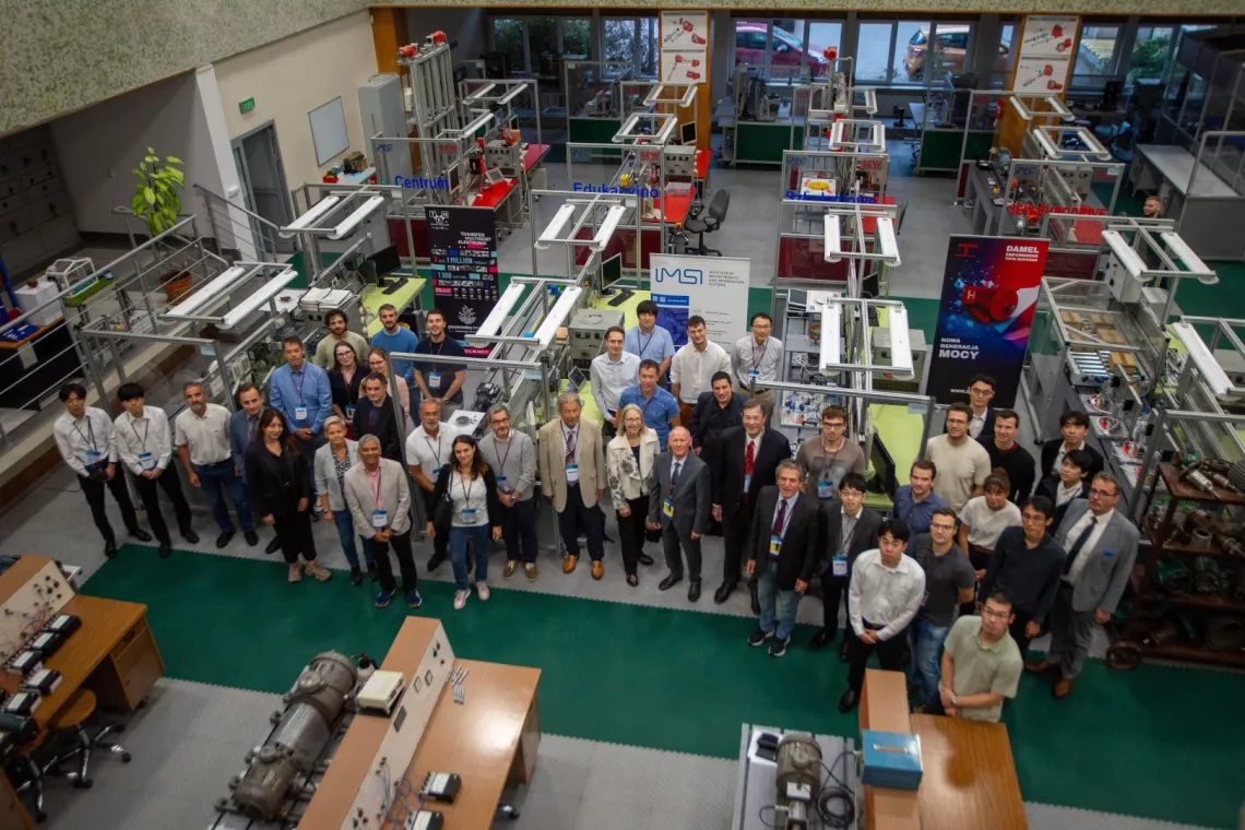 Liczna grupa osób pozująca do zdjęcia w laboratorium, w którym znajduje się wiele różnych stanowisk z maszynami innymi urządzeniami