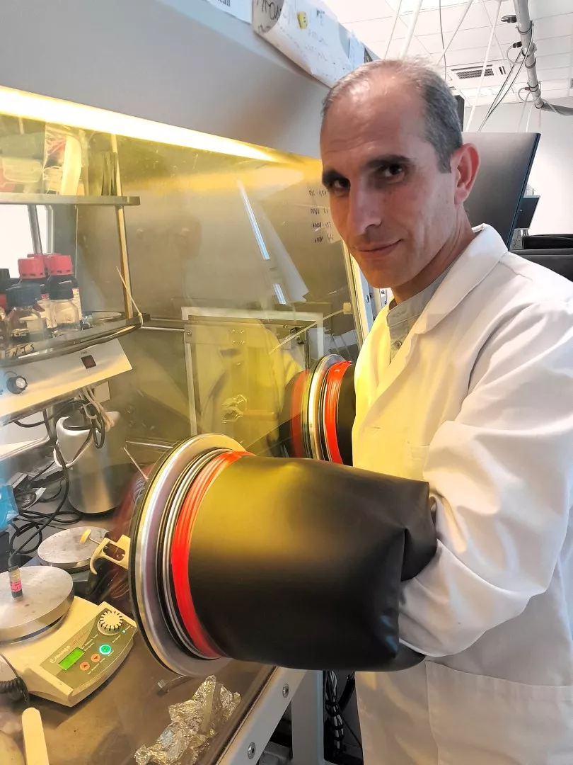 Osoba w fartuchu laboratoryjnym korzysta z rękawicy do manipulowania materiałami wewnątrz zamkniętej przestrzeni na stanowisku w laboratorium. Na stole roboczym widoczne są różne urządzenia laboratoryjne.