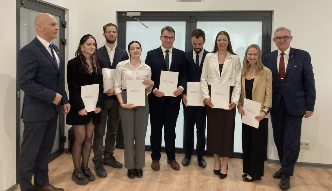 Grupa siedmiu młodych osób trzymających certyfikaty. Są ubrani w formalne stroje i stoją przed szklanymi drzwiami. Towarzyszą im dwaj mężczyżni w garniturach.