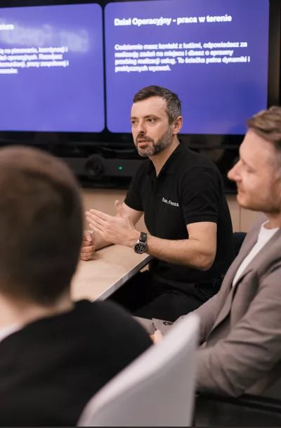 Mężczyzna z krótkim zarostem, w czarnej koszulce z nazwą firmy, siedzi przy stole przemawia do zebranych