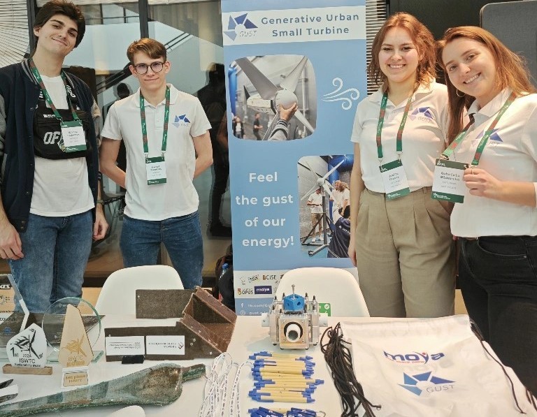 Grupa czterech osób stojąca za stołem na wystawie. Prezentują modele, broszury i materiały promocyjne związane z małymi turbinami miejskimi. W tle stoi baner z napisem 'Generative Urban Small Turbine - Feel the gust of our energy!' 