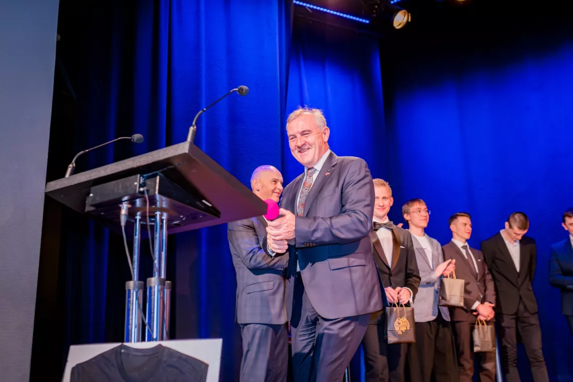 Uśmiechnęty mężczyzna trzyma mikrofon w ręki, z tyłu widać grupę nagrodzonych osób
