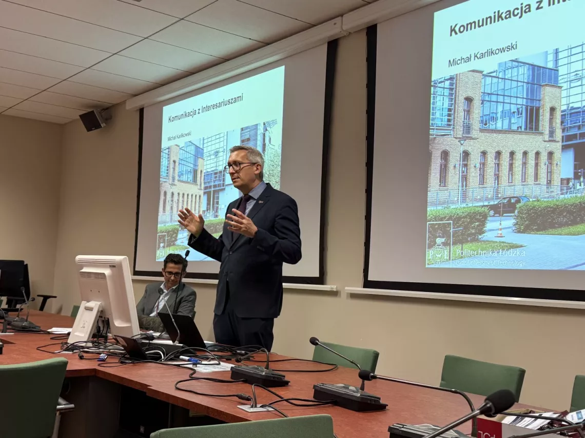 Mężczyzna w graniturze przemawia, stojąc za stołem w sali konferencyjnej. W tle ekran ze slajdem, na którym wyświetla się  napis komunikacja z interesauriuszami, Michał Karlikowski
