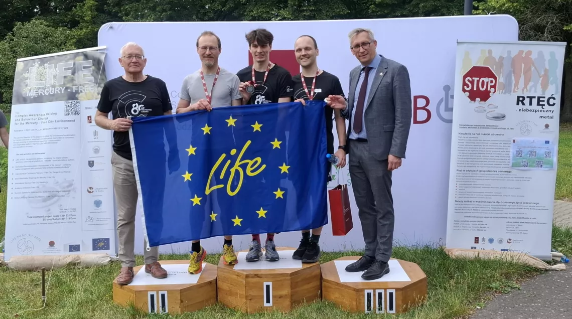 Pięciu mężczyzn, w tym trzech w strojach sportowych i z medalami na szyi. Stoja na podium, trzymając falagę unijną z napisem life