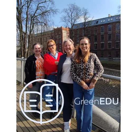 Cztery kobiety stoją na drewnianym moście z metalową barierką. W tle widać budynki z cegły oraz drzewa bez liści