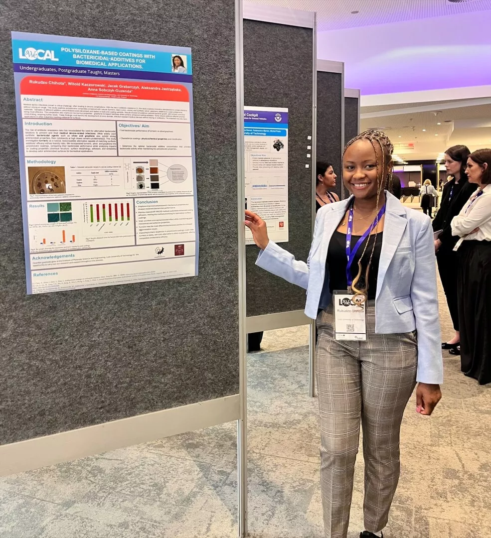 Dziewczyna w niebieskiej marynarce stoi przy ściance, na której jest powieszony naukowy plakat.