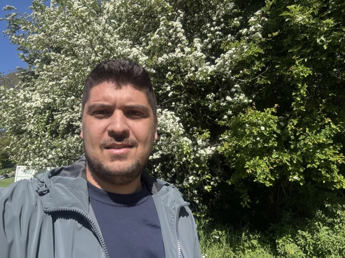 Mężczyzna z "dwudniowym" zarostem w kurtce stoi na tle kwitnącego krzewu