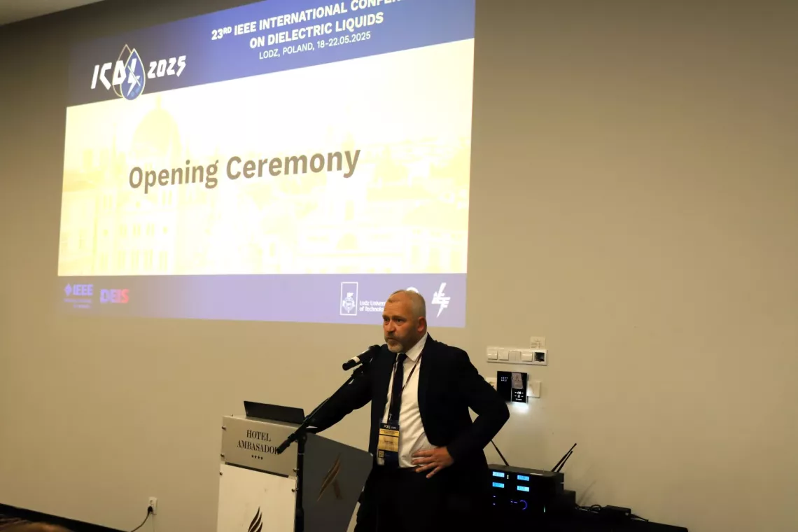 Elegancko ubrany mężczyzna stoi przy mównicy, z tyłu wyświetla się  napis Opening Ceremony