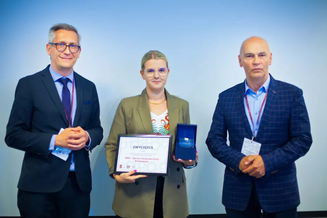 Dwóch mężczyzn w garniturach i kobieta w środku w marynarce trzymająca w dłoniach dyplom i statuetkę