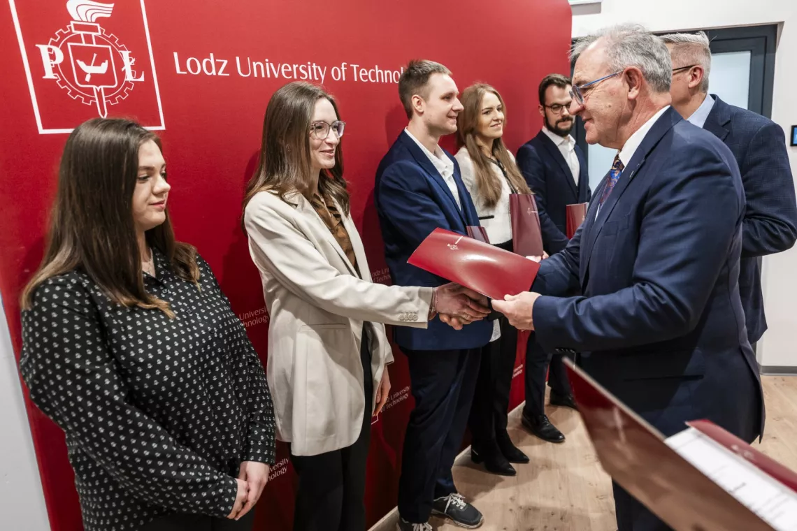 Elegancko ubrany mężczyzna w okularach składa gratulacje młodej kobiecie i wręcza jej teczkę na dokumenty. W szeregu z kobietą stoją jeszcze cztery inne osoby.