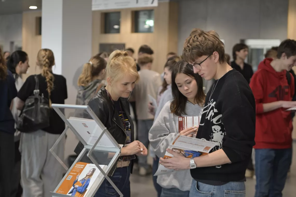 Młodzież w holu budynku, na pierwszym planie dwoje uczniów trzyma informator i rozmawia z młodą kobietą