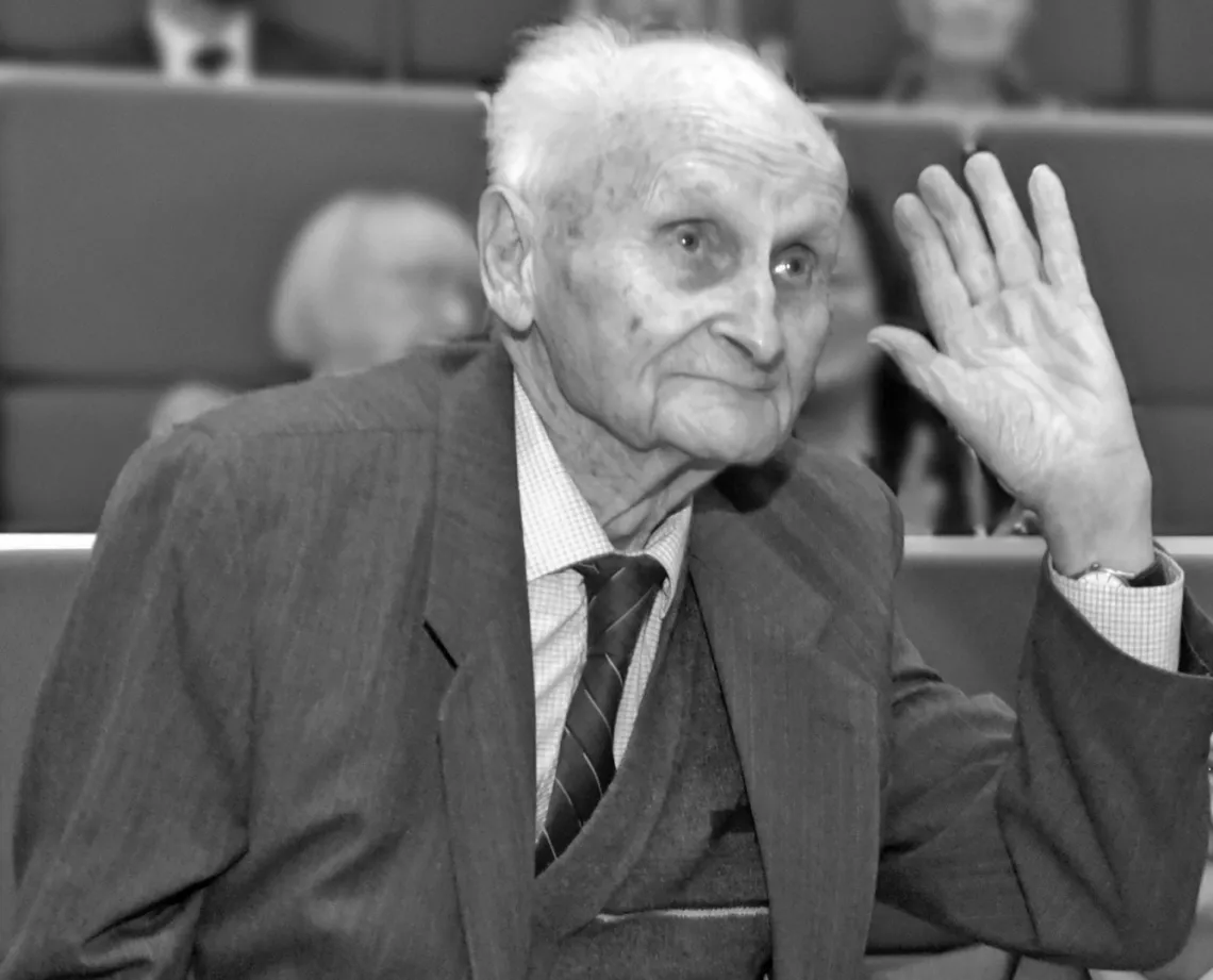 Starszy mężczyzna ubrany w garnitur i krawat, podnoszący rękę na powitanie w obecności innych osób w oficjalnym otoczeniu.