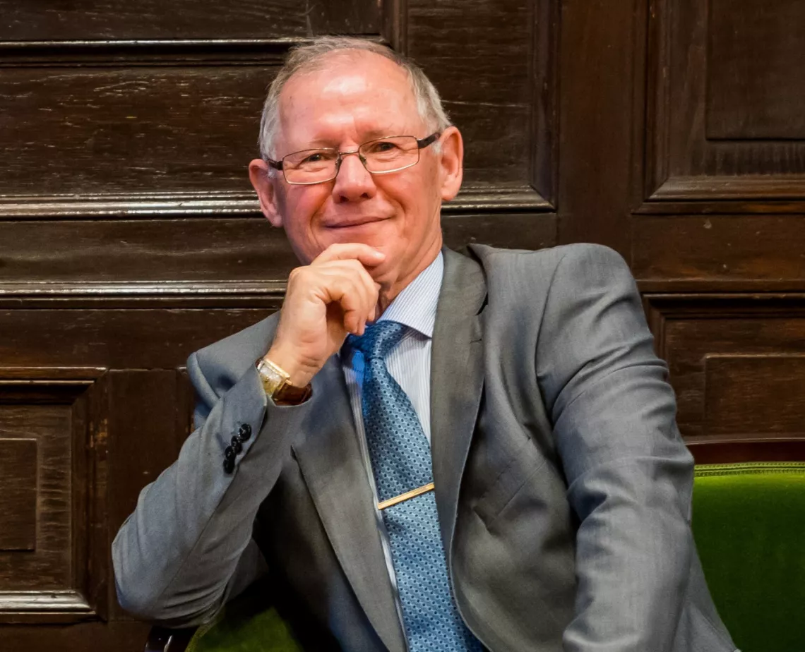 Usmiechnięty mężczyzna w garniturze i w okularach, siedzi przy stole, podpierając dłonią twarz