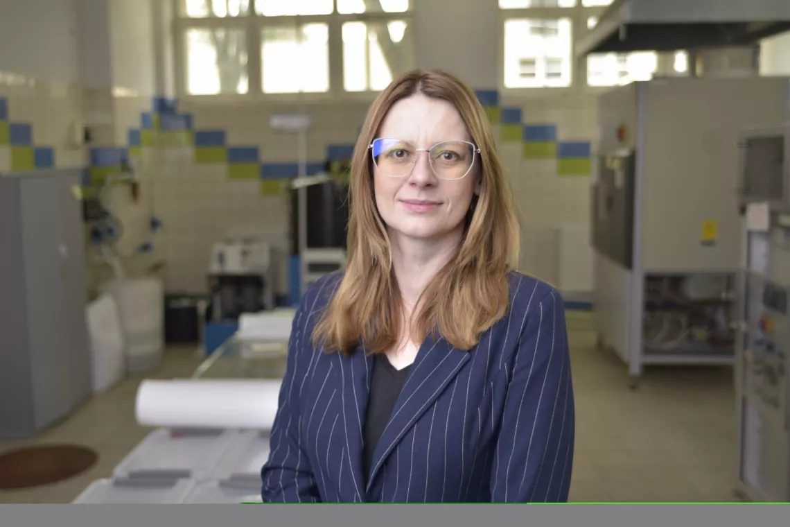 Kobieta z długimi włosami, ubrana w marynarkę w prążki, stoi w laboratorium. W tle widoczne są różne urządzenia oraz ściany wyłożone kafelkami z niebieskimi i zielonymi akcentami. 