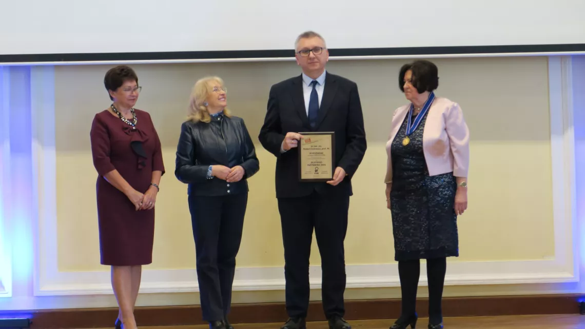 Elegancko ubrany mężczyzna w okularach trzyma w ręku dyplom, towarzyszą mu trzy kobiety także w odświętnych strojach.