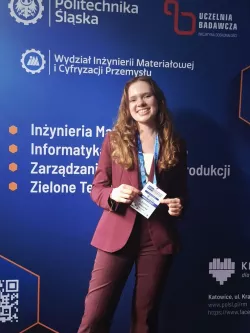 Uśmiechnięta młoda dziewczyna, z długimi jasnymi włosami, ubrana w garnitur i z identyfikatorem w ręku stoi na tle ścianki