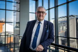 Mężczyzna w formalnym ubraniu stoi na tle opzreszkolen ściany, oparty o poręcż (ujęcie postaci w planie średnim)