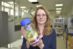 Kobieta znajdująca się w laboratorium trzyma przezroczysty plastikowy słoik z niebieską pokrywką. Wewnątrz słoika znajdują się kolorowe opaski. W tle widoczne są różne urządzenia i półki. 
