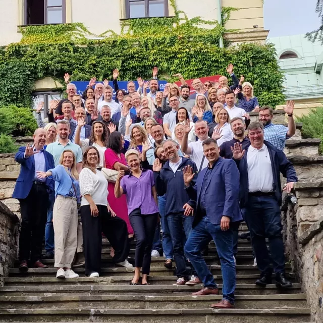 Grupa kilkudziesięciu osób stoi na schodach do budynku, w tle duzo zieleni i fasada, na ktorej widać pnącze