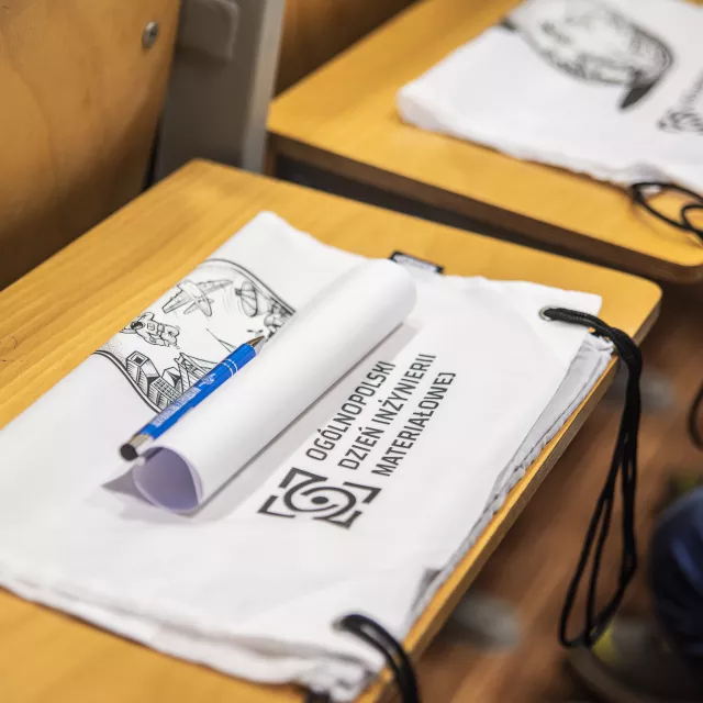 Rząd pulpitów z białymi torbami z nadrukowanym tekstem OGÓLNOPOLSKI DZIEŃ INŻYNIERII MATERIAŁOWEJ i logo. Na jednym z blatów widoczne są również zwinięte papiery i niebieski długopis.