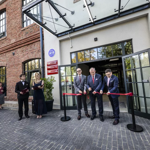 Przecięcie wstęgi przez trzech mężczyzn, na tle zrewitalizowanego budynku z czerwonej cegły. Obok stoi para młodych, eleganko ubranych ludzi prowadzących uroczystość oraz grupa gości.