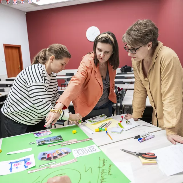 Trzy kobiety pracują nad planszą, która leży na stole. Obok leżą nozyczki, flamastry i różne materiały papierowe