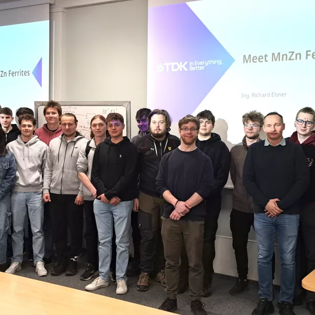 Studenci stoja na tle ekranów, na których wyświetla się napis Meet MnZn Ferrites
