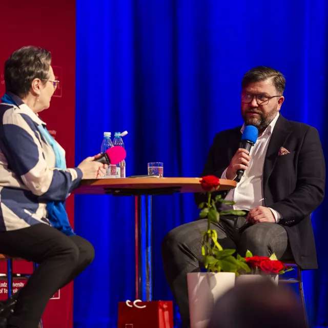 Dwie osoby przy okragłym stole prowadzą rozmowę. Obie trzymają mikrof w dłoniach.