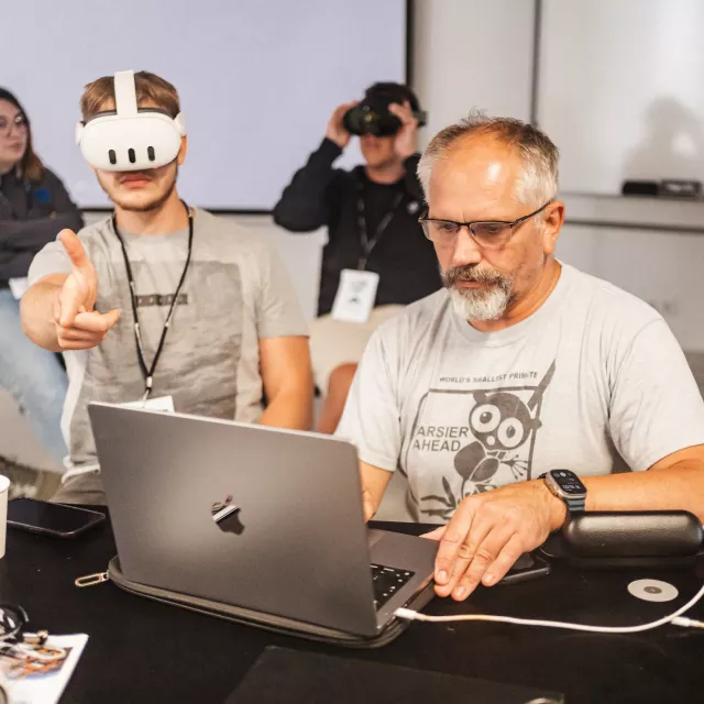 Mężczyzna pracuje przy laptopie, obok student z goglami VR