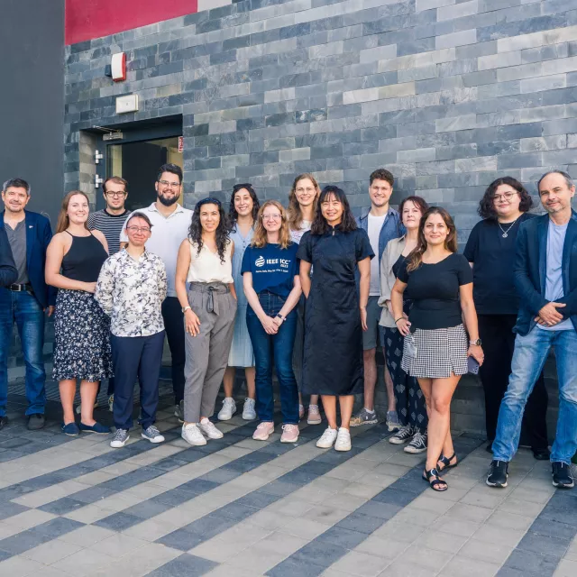 Grupa 17 osob pozuje do zdjęcia na tle ściany budynku, wułożonej szarymi płytkami