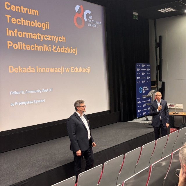 Dóch mężczyzn stoi przed publicznością siedzącą na widowni, jeden z nich przemawia do mikrofonu