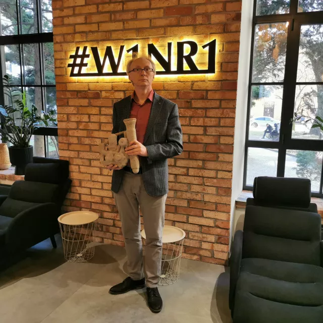 Mężczyzna w okularach , szarej marynarce i kolorowej koszuli, trzyma w rękach odlew. Z tyłu ściana z cegły z napisem #W1NR1 i okna