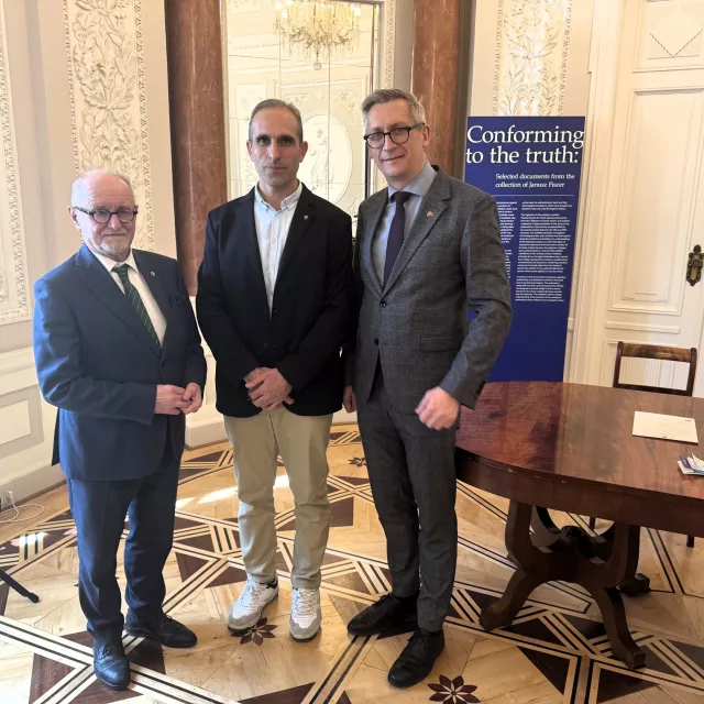 Trzy osoby stoją w pokoju z ozdobnym wystrojem. Są ubrani w formalne stroje. Za nimi znajduje się baner z napisem "Conforming to the truth: The future of science and technology." Po ich prawej stronie znajduje się drewniany stół z kilkoma papierami.