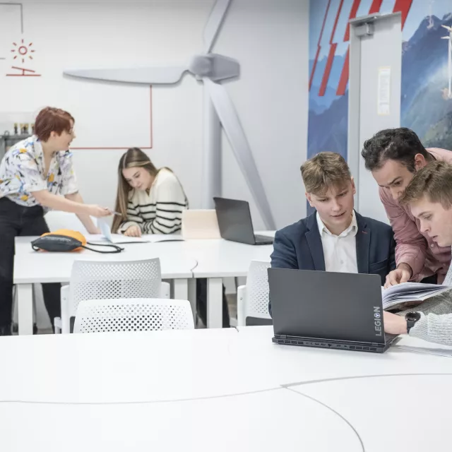 Studenci w czasie zajęć pracują przy laptopach, konsultująć się z prowadzącymi zajęcia